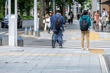 Urban street scene of featuring morning commuters. A cyclist and pedestrian, both wearing backpacks, wait for traffic signal at a crosswalk, as other commuters go about their daily business.