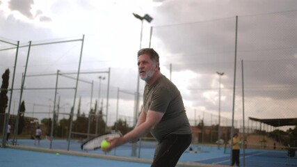 Mature tennis player serving powerfully on blue court, displaying athletic skill and dedication under cloudy sky while practicing recreational sport