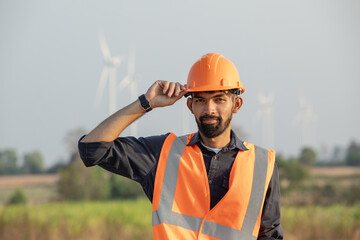 Handsome bearded young engineer wearing a hard hat and vest standing on wind farm.