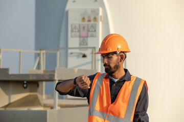 Handsome bearded young engineer wearing hard hat and vest looking at wristwatch while standing on wind farm