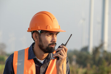 Young bearded engineer or maintenance technician wearing a hard hat using walkie-talkie on a wind farm.
