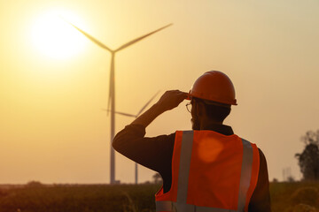 Young male engineer in hard hat and vest standing on wind farm during sunset background.