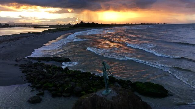 Sunset waves crash against a rocky outcrop, spray rising around a lone statue silhouetted in gold&mdash;still and solemn amid the ocean&rsquo;s restless glow.