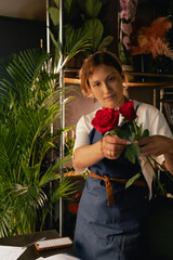 Middle-aged female florist in apron arranging fresh flowers for bouquet in the flower shop. Small...