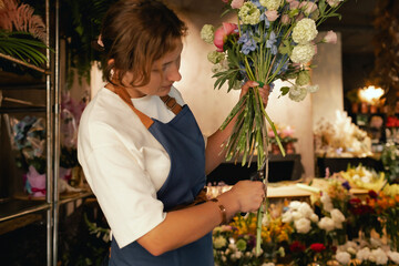 Middle-aged female florist in apron arranging fresh flowers for bouquet in the flower shop. Small...