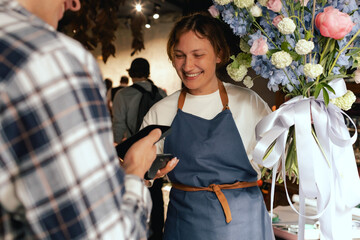 Flower shop: Female florist in apron selling bouquet of flowers, customer paying with smartphone...