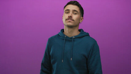 Obraz premium Young hispanic man in teal hoodie stands against vibrant purple background, exuding casual style and confidence in an isolated setting.