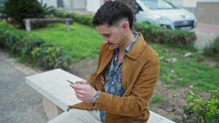 Young hispanic man wearing jacket captures selfie on smartphone in outdoor park surrounded by greenery and benches on a calm day
