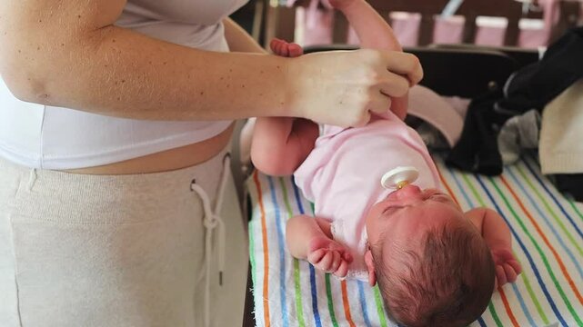 Close-up video shows a mother gently dressing her newborn baby in a white bodysuit after a diaper change on a colorful mat. The scene emphasizes parental care and the routine of early infancy
