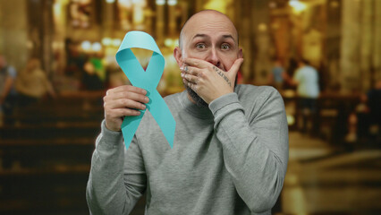 Man holding cancer awareness ribbon in church setting, exhibiting emotional response with...