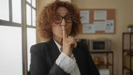 Hispanic woman in black blazer gestures for silence in a bright office, standing confidently by a...