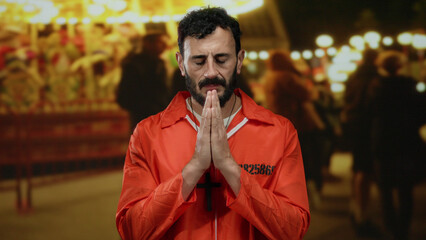 Hispanic man in orange prison jumpsuit with beard prays at a fairground carousel, juxtaposing...