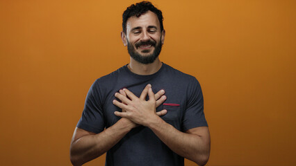 Bearded hispanic man smiling warmly against a vivid orange background with hands on heart symbolizing gratitude and positivity