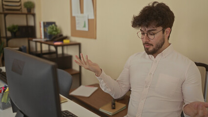 Young man with beard working at computer in office room, looking thoughtful and engaged, surrounded by books and plants, creating a casual yet professional setting indoors.