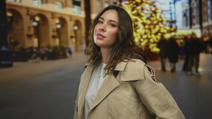 Fototapeta premium Woman in trenchcoat stands confidently on city street adorned with festive lights, radiating a warm expression in an urban setting during the holiday season.