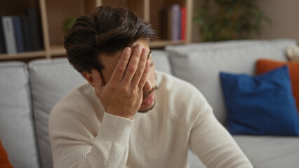 Young hispanic man looking stressed in cozy living room with hand on face, wearing casual sweater, surrounded by modern decor and colorful cushions on the couch.