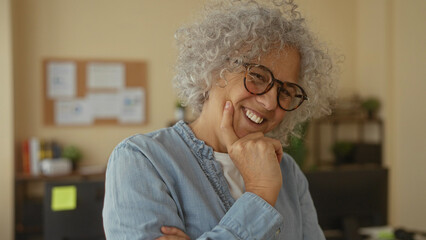 Woman smiling in office with grey hair and glasses, surrounded by books and plants in a relaxed...