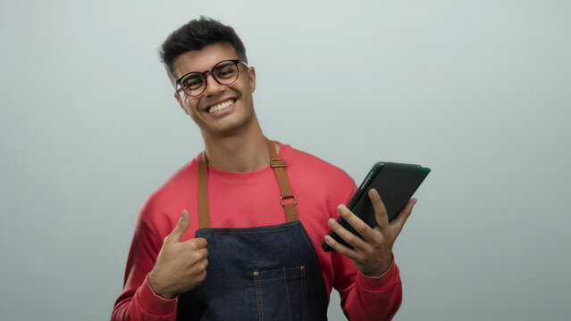 Young man wearing glasses and apron smiles while holding tablet and giving thumbs up against white background