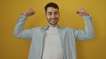 Young hispanic man smiling confidently flexing muscles on an isolated yellow background showcasing...