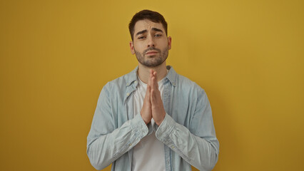 Young hispanic man with hands clasped in a hopeful gesture against a vibrant yellow background, portraying an attractive and contemplative expression in a casual setting.
