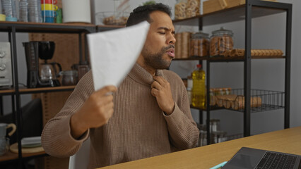 Young man in office using paper to cool down while feeling warm indoors with shelves and kitchen...