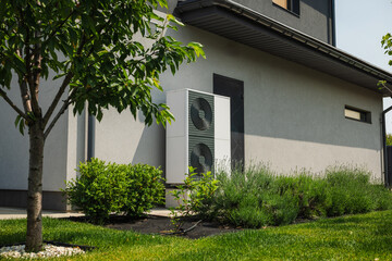 Fototapeta na wymiar Modern heat pump unit installed next to a residential house wall, surrounded by green lawn, bushes, and young trees in a landscaped yard