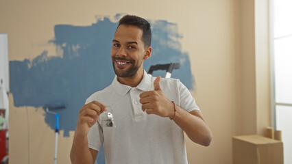Young man giving thumbs up in new home, holding keys, signifying successful move, with painted...