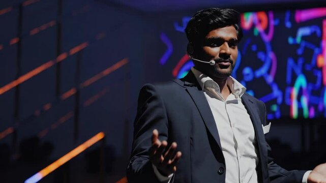 Indian businessman wearing suit and headset microphone presenting on stage at a corporate business conference with colorful graphic on big screen in the background, inspiring his audience