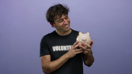 Hispanic man with a volunteer shirt smiling while holding a piggy bank against a purple background,...