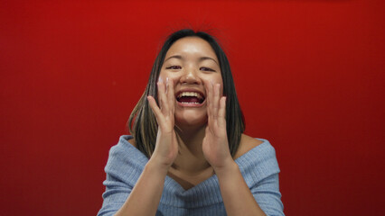 Woman shouting against a vibrant red wall background, expressing enthusiasm with hands near face, wearing casual blue sweater, isolated from chinese culture.
