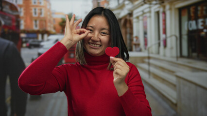 Woman smiling holding heart paper making ok gesture on city street with red bus in background...