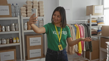 Woman smiling during videocall in donations center surrounded by clothing and food items wearing green volunteer shirt in indoor room setting suggesting charitable activity.