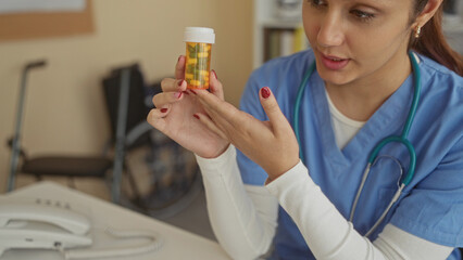 Hispanic woman doctor in hospital room holding medication bottle, wearing scrubs, examining pills with focus, stethoscope around neck, office phone in background.