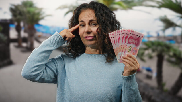 Middle-aged woman at seaside holding chinese yuan with a thoughtful gesture, symbolizing financial contemplation by the beach.