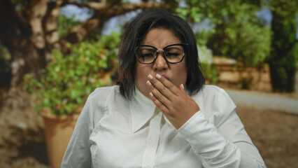 Woman in a park expressing disgust with hand covering mouth, wearing glasses and white shirt,...