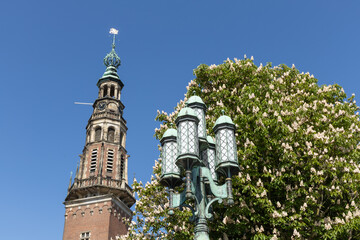 The ornate City Hall tower of Leiden rises grandly next to a blossoming tree and an antique streetlamp under a vibrant blue sky.