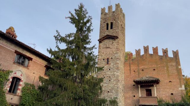 Historic clock tower and medieval architecture in Grazzano Visconti, Italy, with ivy covered walls and a central evergreen tree under clear sky