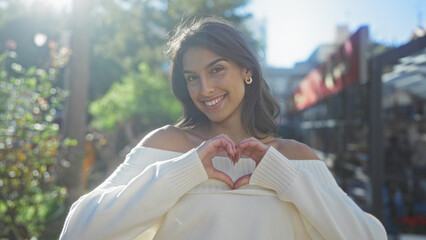 Woman smiling in city park with sunlight highlighting her features, wearing off-shoulder sweater, making heart shape with hands, displaying joy and beauty outdoors.