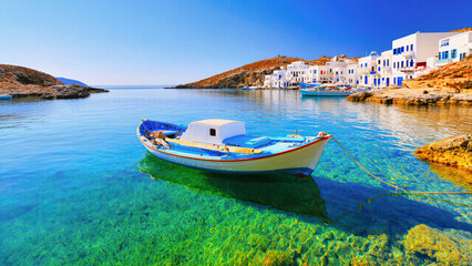 Obraz premium A small traditional boat floats gently on the crystal-clear turquoise sea near whitewashed coastal houses under a bright blue sky on a peaceful sunny day.