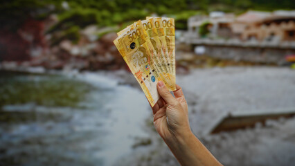 Hand holding philippine pesos outdoors by the seaside, depicting investment and business themes in a scenic town setting.