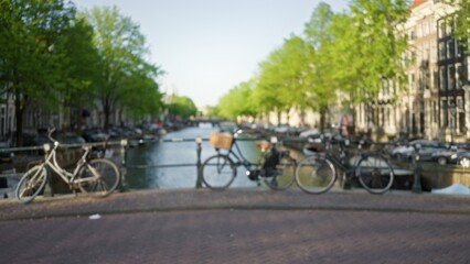 Blurred bicycles rest against a fence with a picturesque amsterdam canal and lush green trees in the background, capturing the serene essence of the netherlands.