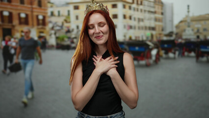 Obraz premium Woman smiling on city street wearing crown with hands on chest surrounded by urban backdrop featuring buildings and people in summer outdoor setting