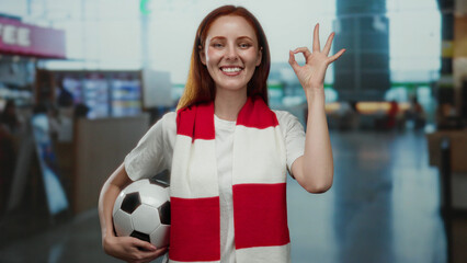 Woman in airport terminal holding soccer ball with red scarf smiles confidently indoors capturing joyful travel spirit and passion for sports showing positive gesture.