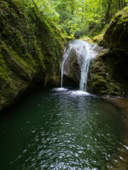 Kazancheto waterfall in Strandzha Nature Park, Bulgaria. Twin streams cascade over mossy rocks into a dark pool, surrounded by lush green forest. Peaceful hidden natural gem in summer