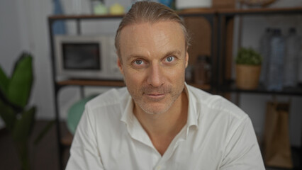 Obraz premium Middle-aged caucasian man with blonde hair in an office setting, wearing a white shirt, exhibiting a confident expression, and surrounded by indoor plants and office items.