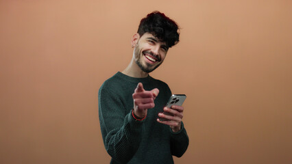Young man with phone smiling and pointing at camera over brown background, conveying connection and engagement with audience, showcasing charm and technology use.