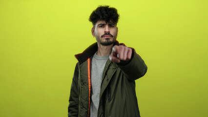 Fototapeta premium Young man pointing with confidence against a bright yellow background, wearing a stylish green jacket, exuding a charismatic and assertive demeanor.
