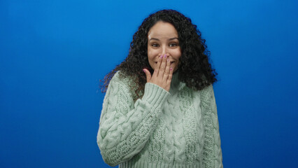 Woman smiling with hand over mouth against vibrant blue background wearing green sweater and curly...