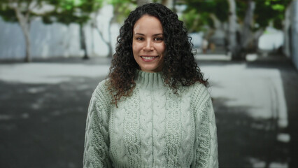 Woman wearing a light sweater smiles outdoors on an urban street surrounded by blurred greenery and trees.