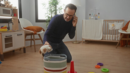 Man organizing toys in cozy bedroom while talking on phone, surrounded by colorful baby items in modern home environment.